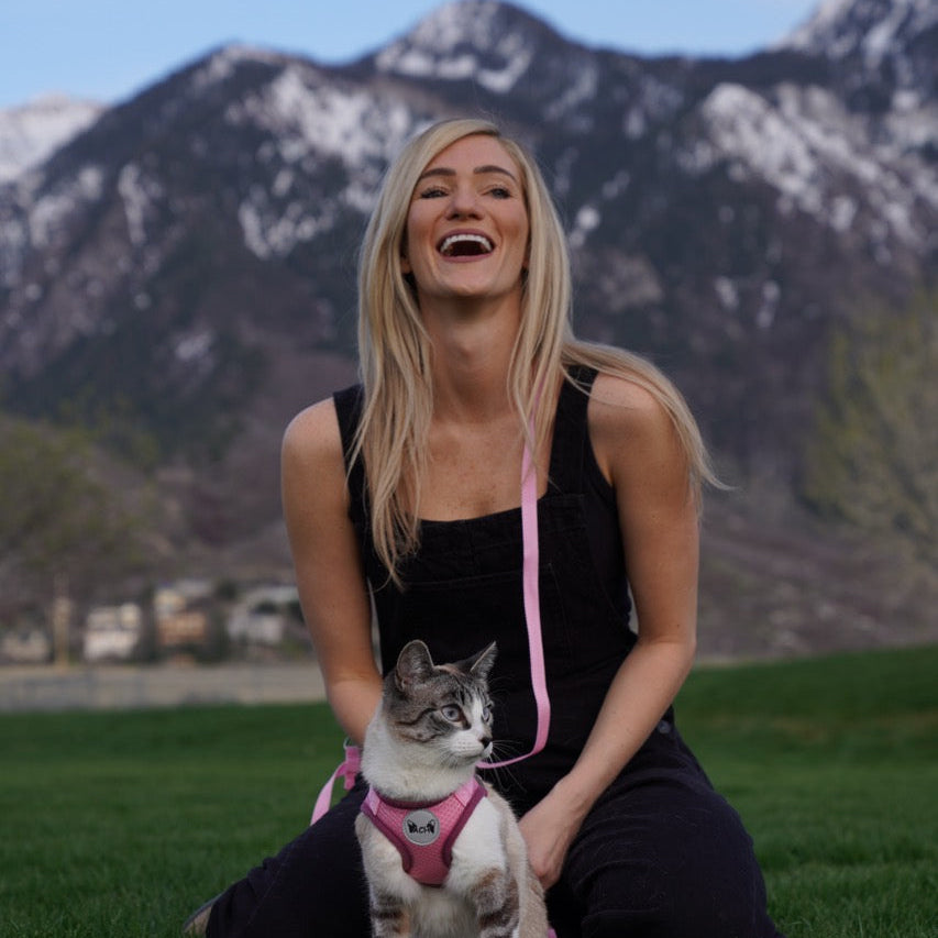 Woman sitting on grass with a cat wearing a pink harness, mountains in the background