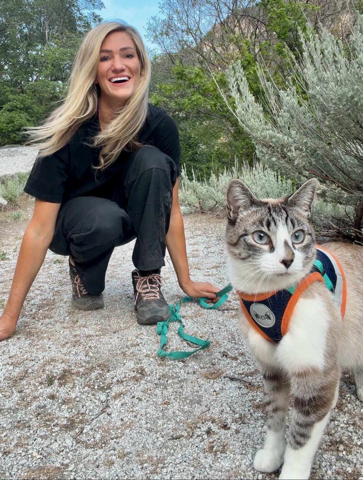 Woman kneeling on a gravel path with a cat wearing a harness, surrounded by greenery.